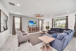 Relax in this spacious living room with a large flat-screen TV, comfortable seating area, and large window with blinds.
