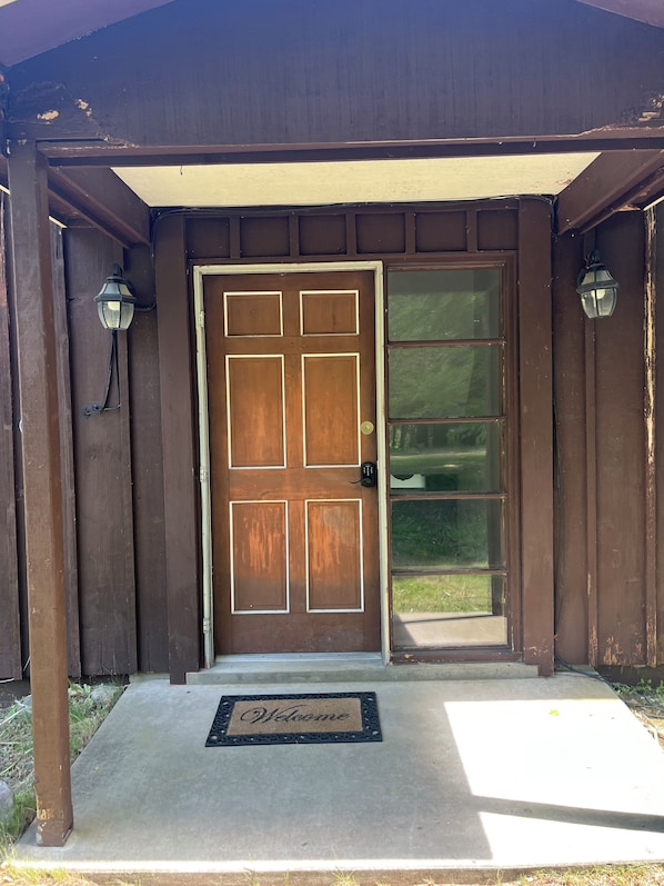 A wooden front door with a glass panel, a welcome mat, and two wall-mounted lanterns.