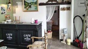 Una habitación acogedora con una zona de bar de estilo antiguo que dice 'CAFÉ DE PARÍS', una pared de piedra rústica, un cuadro que representa una escena de río y un umbral que da a otra habitación.