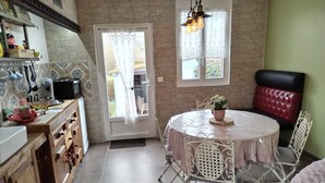 Un comedor con una mesa redonda, sillas blancas y una silla con cojín rojo. Hay una planta en maceta sobre la mesa, y se ve una ventana con cortinas.