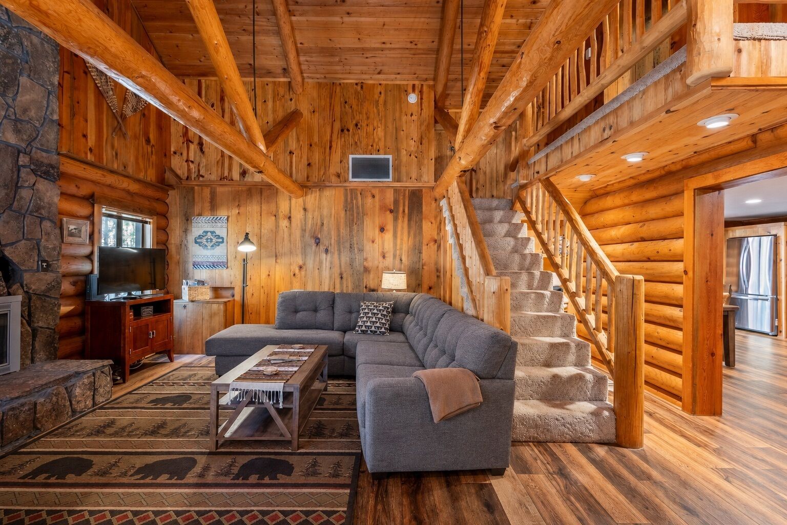 A log cabin interior with a stone fireplace, a sectional sofa, a television, and a staircase.