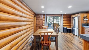 Een houten blokhut vanbinnen met een houten eettafel, stoelen, een koelkast en een raam met uitzicht op bomen.