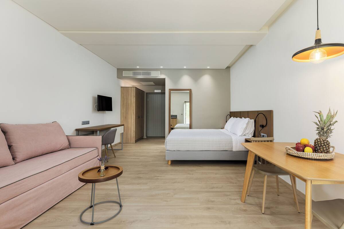 Ein modernes Hotelzimmer mit einem Bett, einem Holztisch mit Obst, einem kleinen runden Tisch mit einer Vase, einer Couch und einem Fernseher an der Wand.