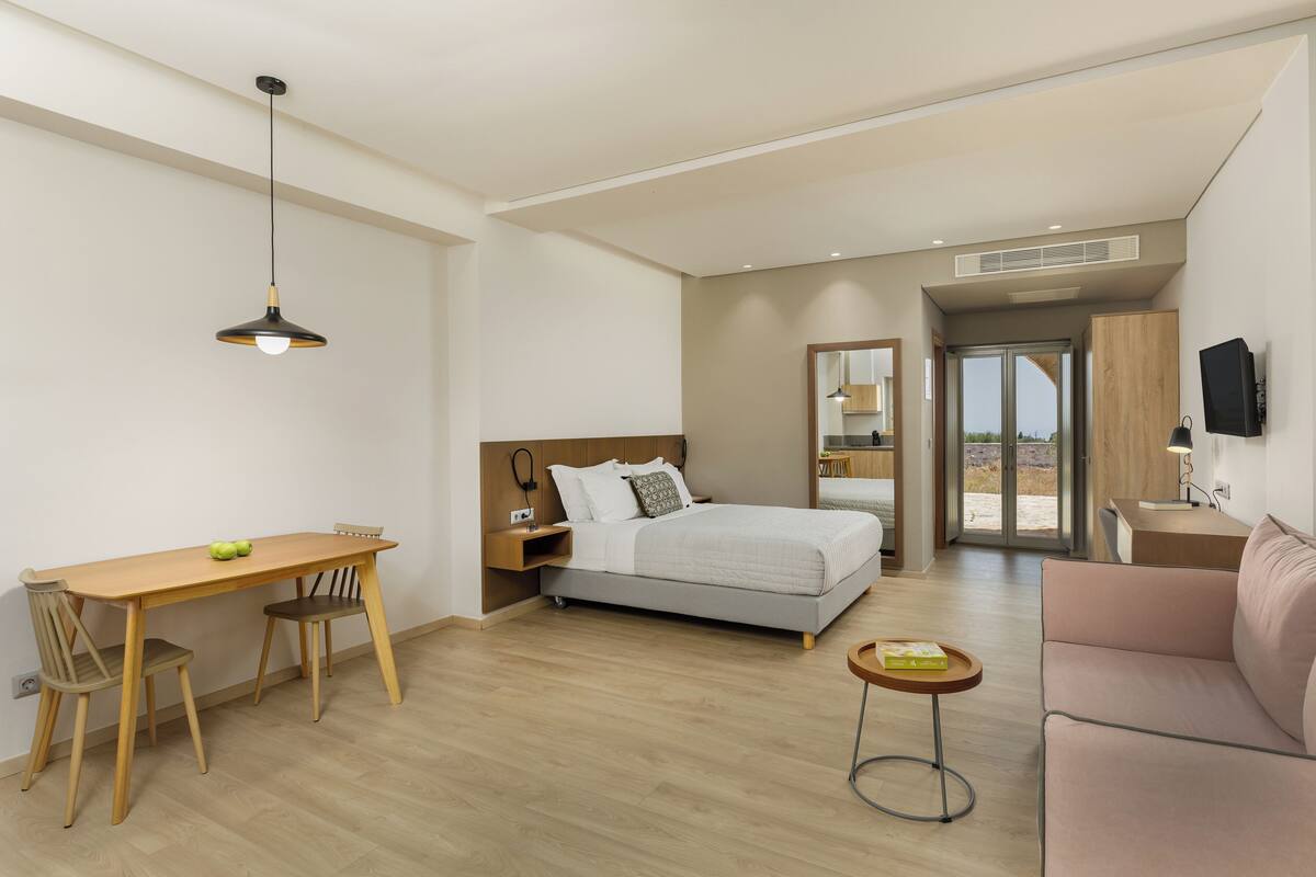 Ein modernes Hotelzimmer mit einem Bett, einem Schreibtisch mit Stühlen, einem kleinen Tisch, einer Couch und Blick ins Freie.