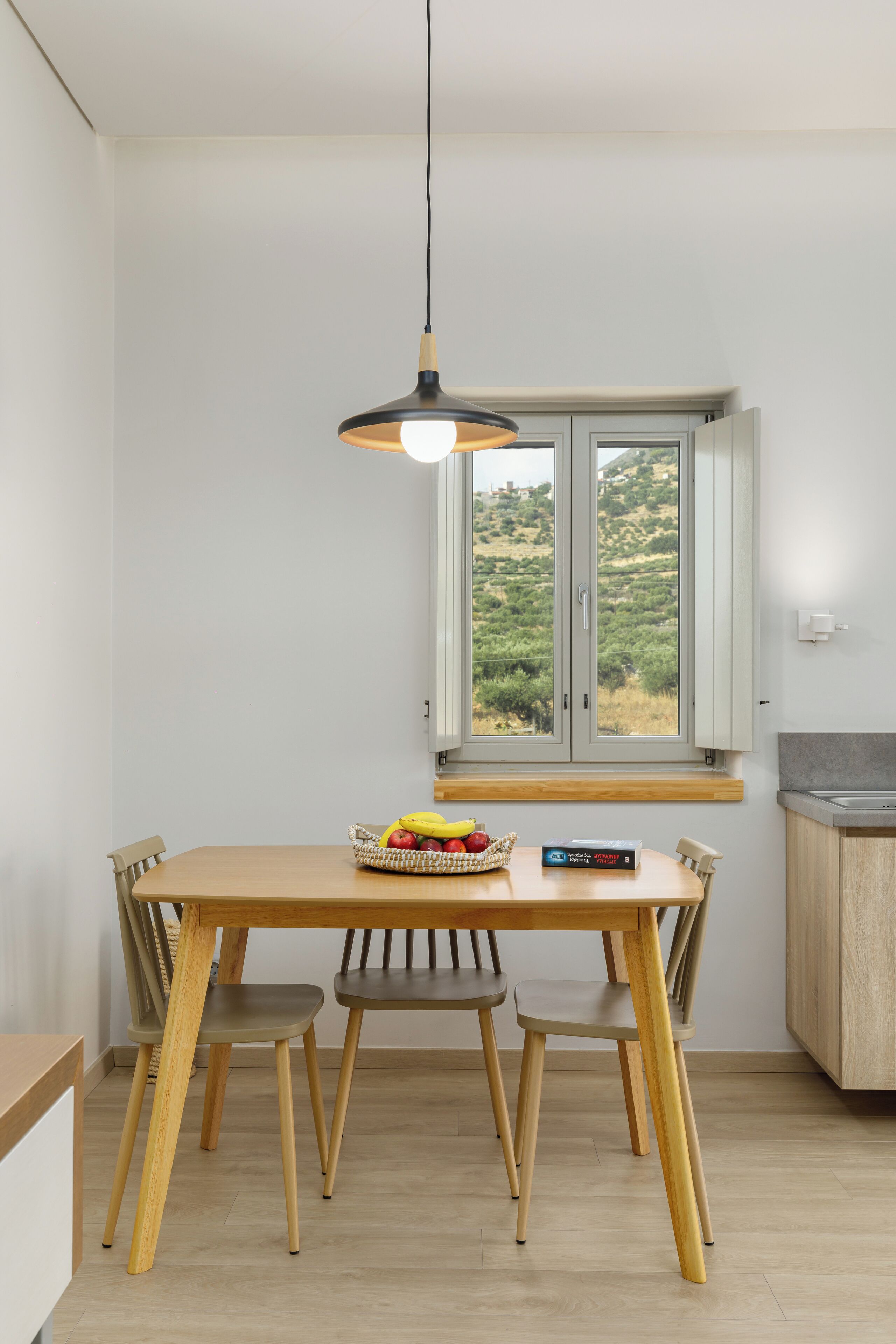Un comedor con mesa de madera, cuatro sillas, una lámpara colgante, ventana con vista a follaje y una cesta de fruta.