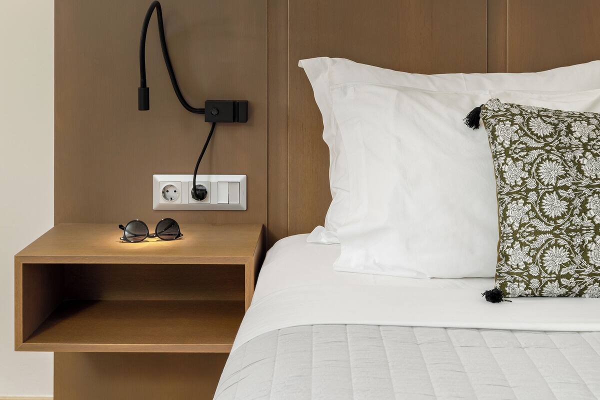 Ein ordentlich bezogenes Bett mit weißer Bettwäsche und einem gemusterten Kopfkissen, ein Nachttisch mit Steckdose und Gläsern sowie eine wandmontierte Ladestation.