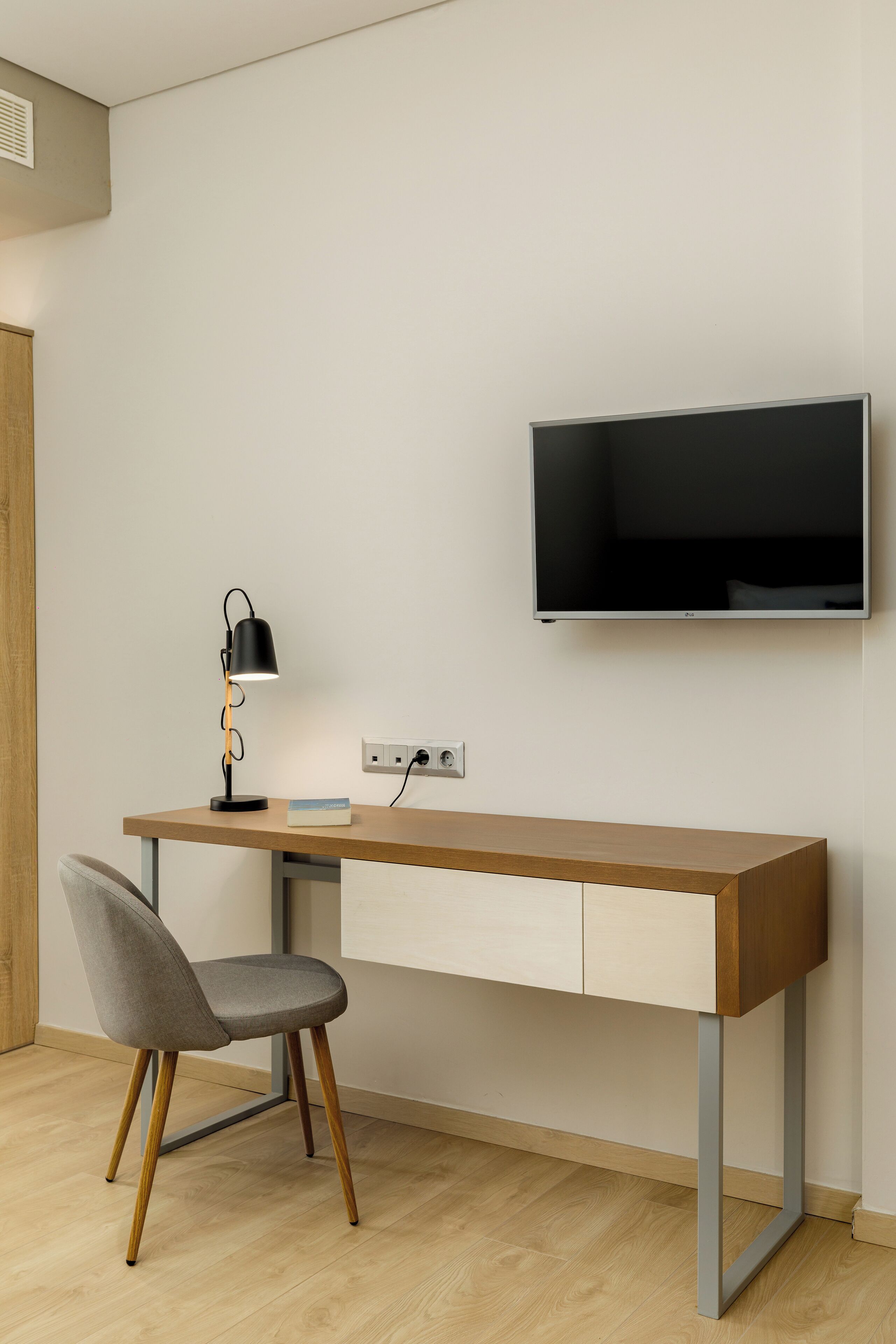 Een moderne kamer met een houten bureau, een grijze stoel, een zwarte bureaulamp en een tv aan de wand.