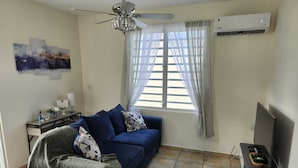 A living room with a blue sofa, a TV stand, a ceiling fan, and a window with curtains. - Cozy Island Home (Quebradillas)