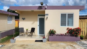 A single-story house with a beige exterior, a grey door, and a window with horizontal blinds. There is a small porch with two chairs and a potted plant. - Cozy Island Home (Quebradillas)