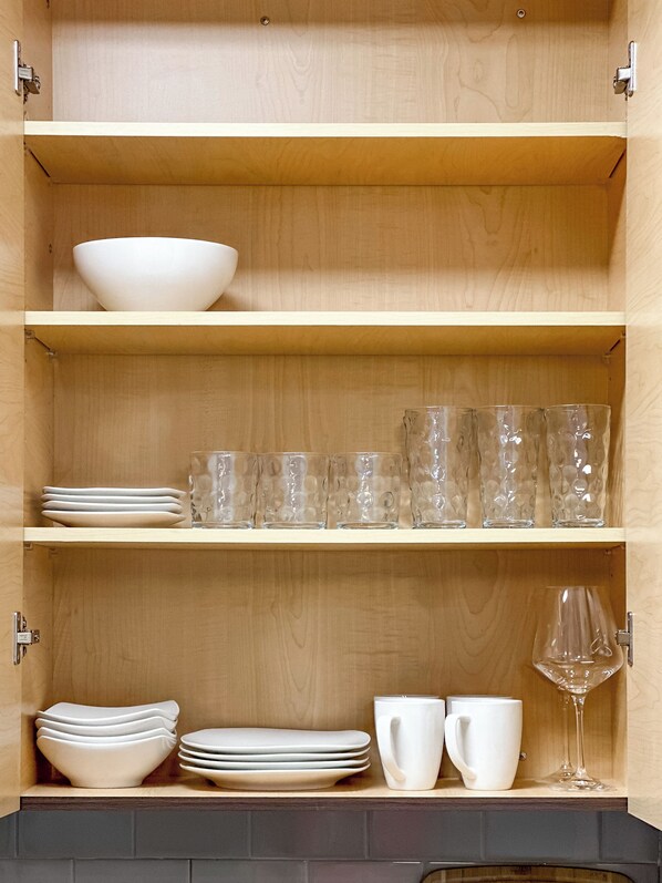 A wooden cabinet with three shelves, each holding different items: a white bowl on the top shelf, stacked plates and clear glasses on the middle shelf, and stacked bowls, plates, white cups, and a wine glass on the bottom shelf.