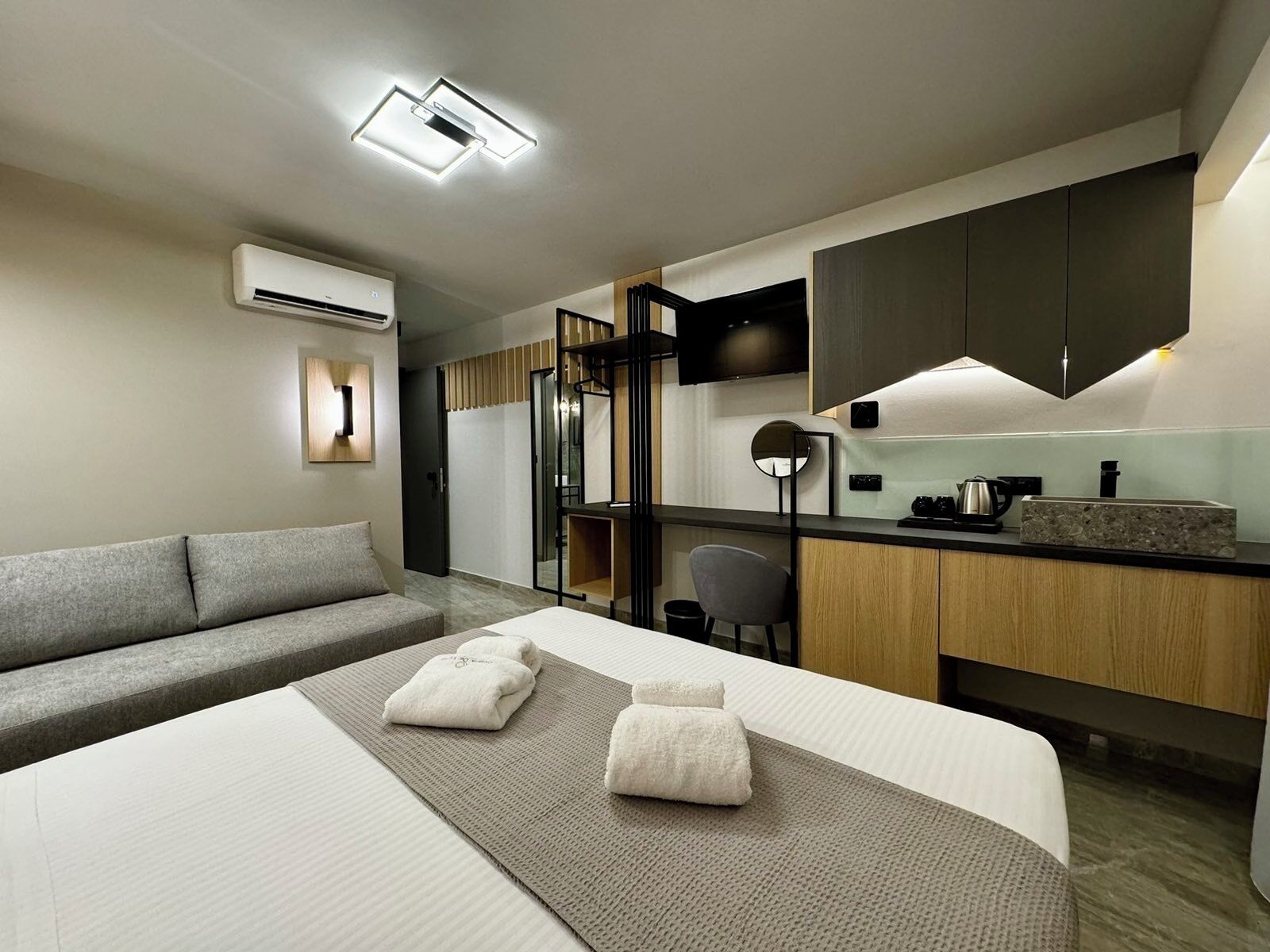 Una habitación de hotel moderna con sofá, cama con toallas, kitchenette y televisor de pantalla plana.