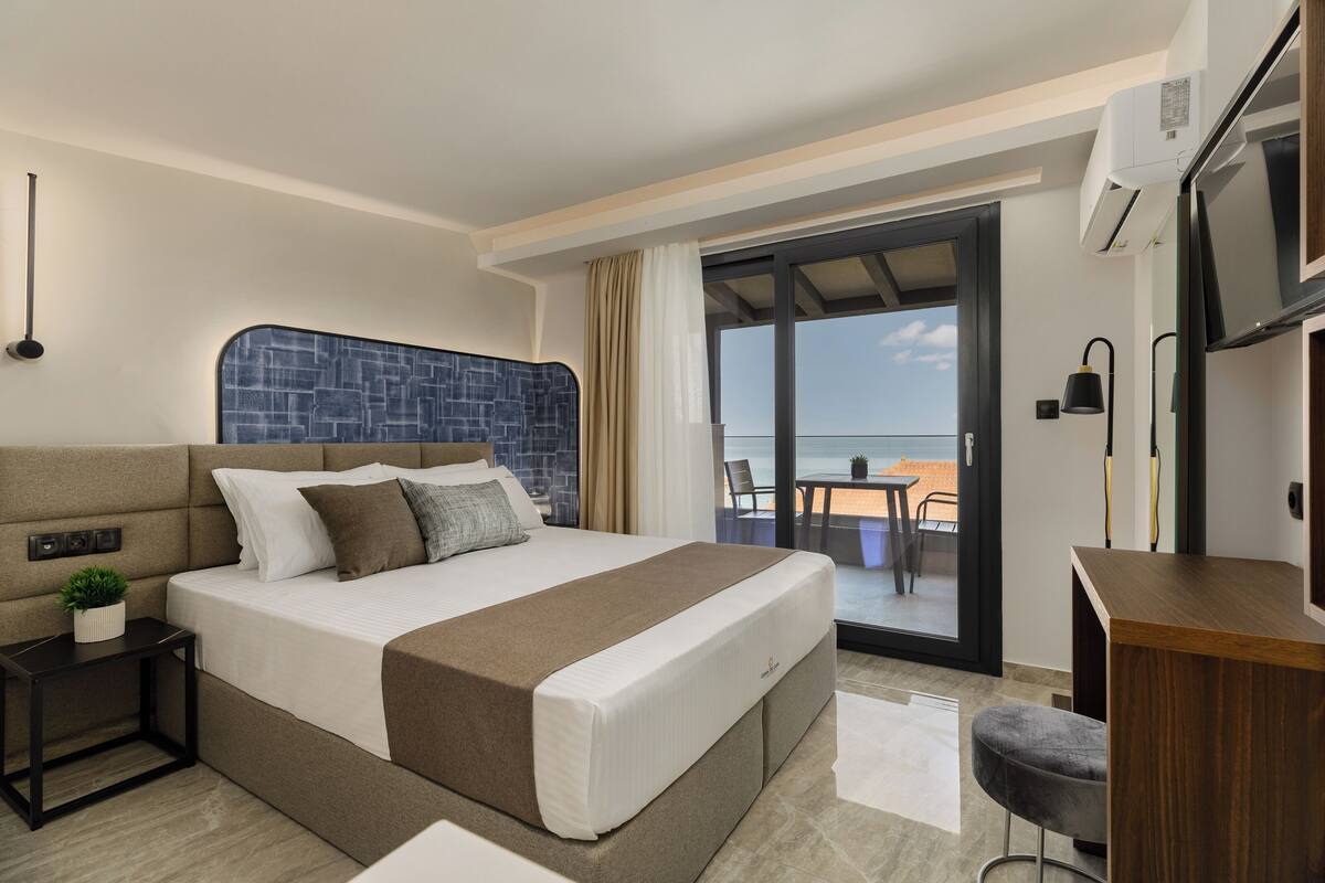 A modern hotel room with a large bed, a small desk, a balcony with a table and chairs, and a view of the sea.