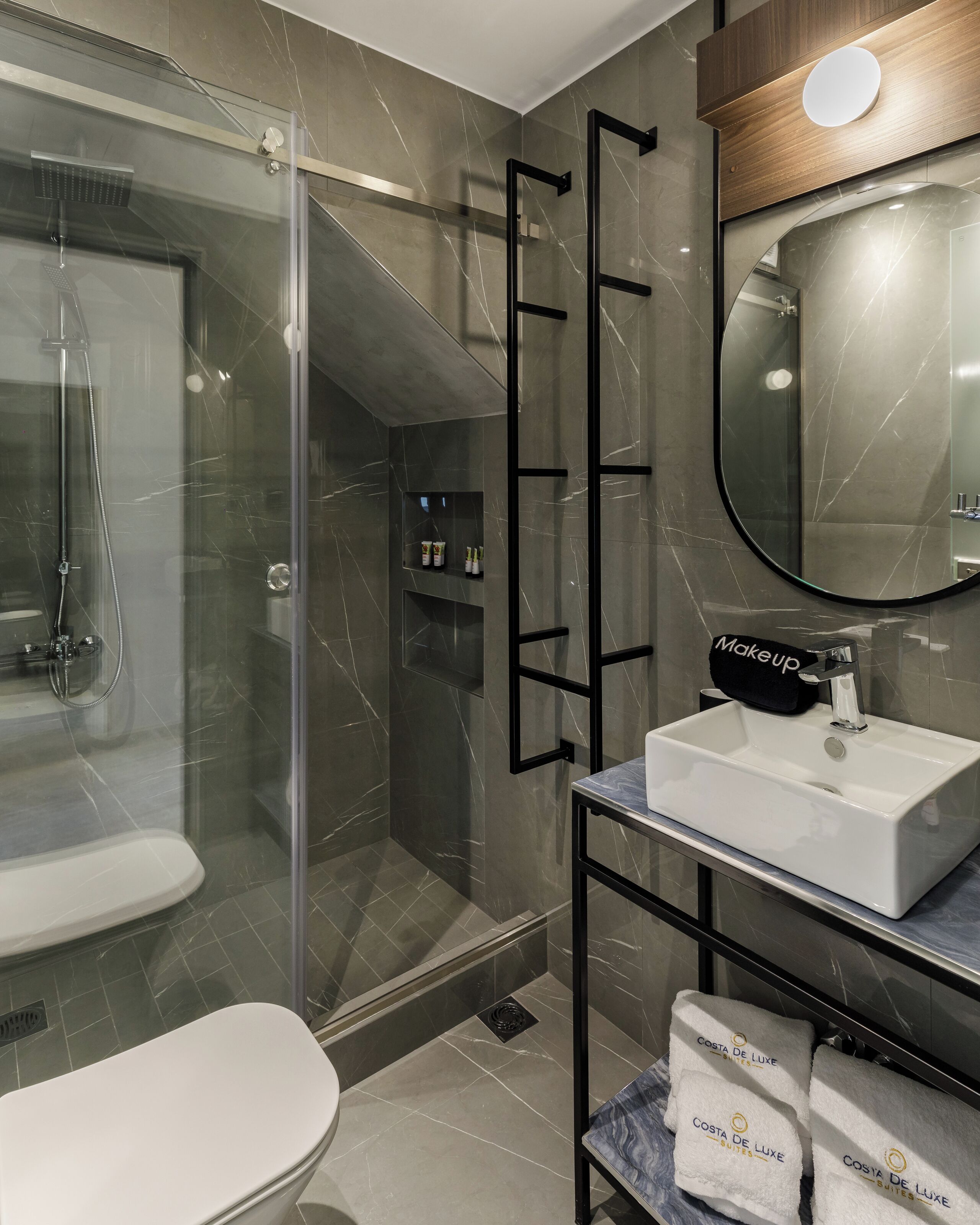 A modern bathroom with a glass-enclosed shower, a white sink, and a mirror above it. There are towels with "Costa de Luxe" branding on the counter.