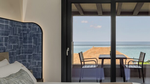 A bedroom with a bed, a view of the beach, and a balcony with a table and chairs. - Costa De Luxe Suites (Dio-Olympos)
