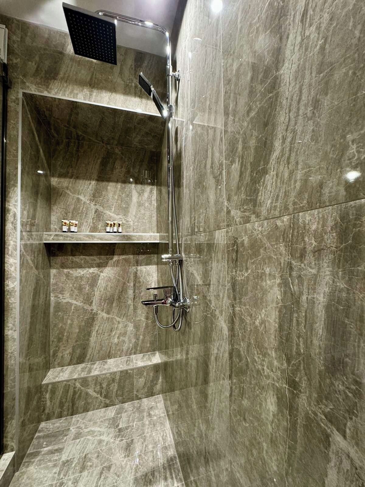 A modern bathroom with a shower, marble walls, and a shelf with toiletries.