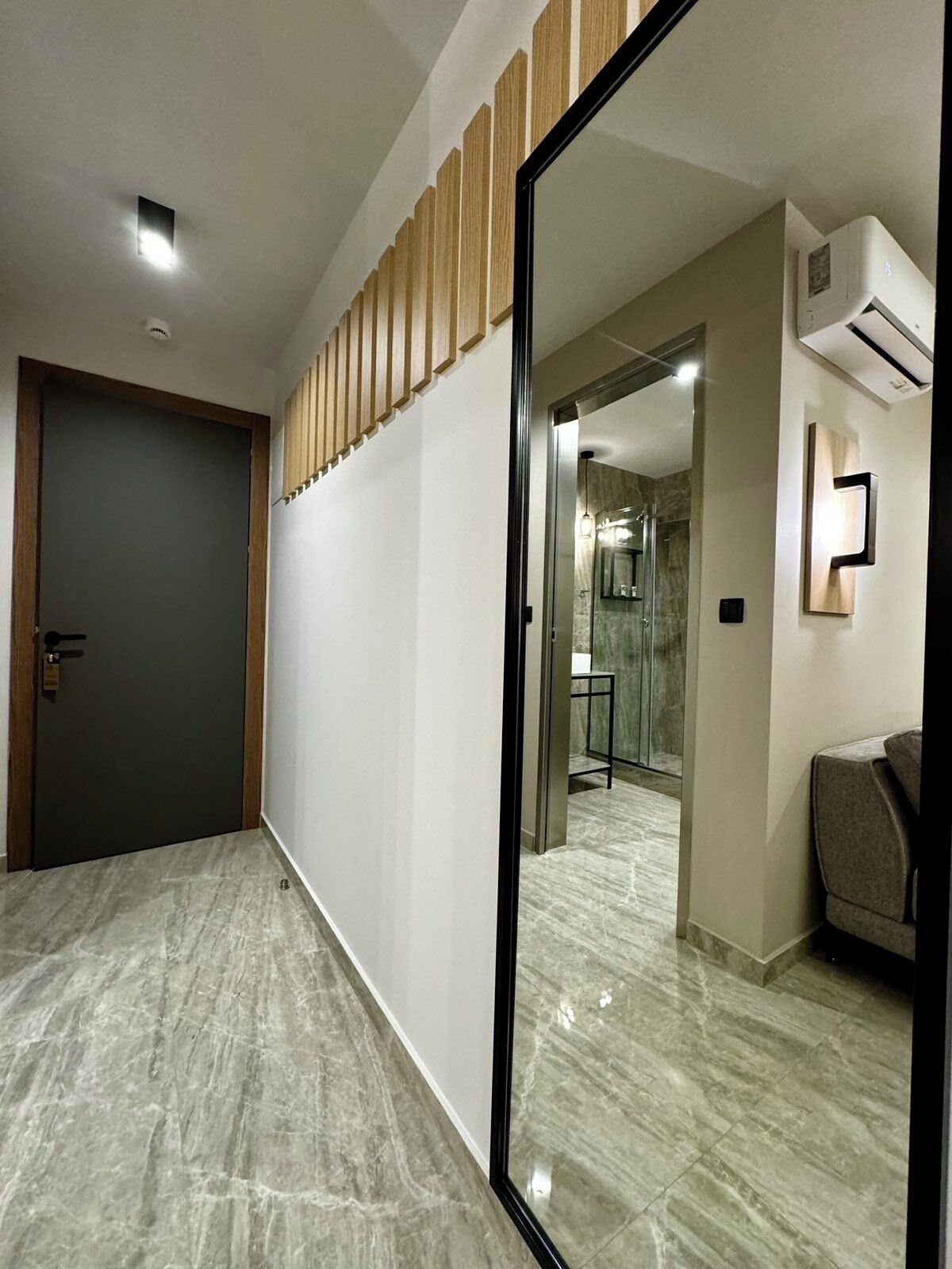 A hallway with a dark door, a wooden panel wall, and a mirrored wall reflecting a bathroom with a marble floor.
