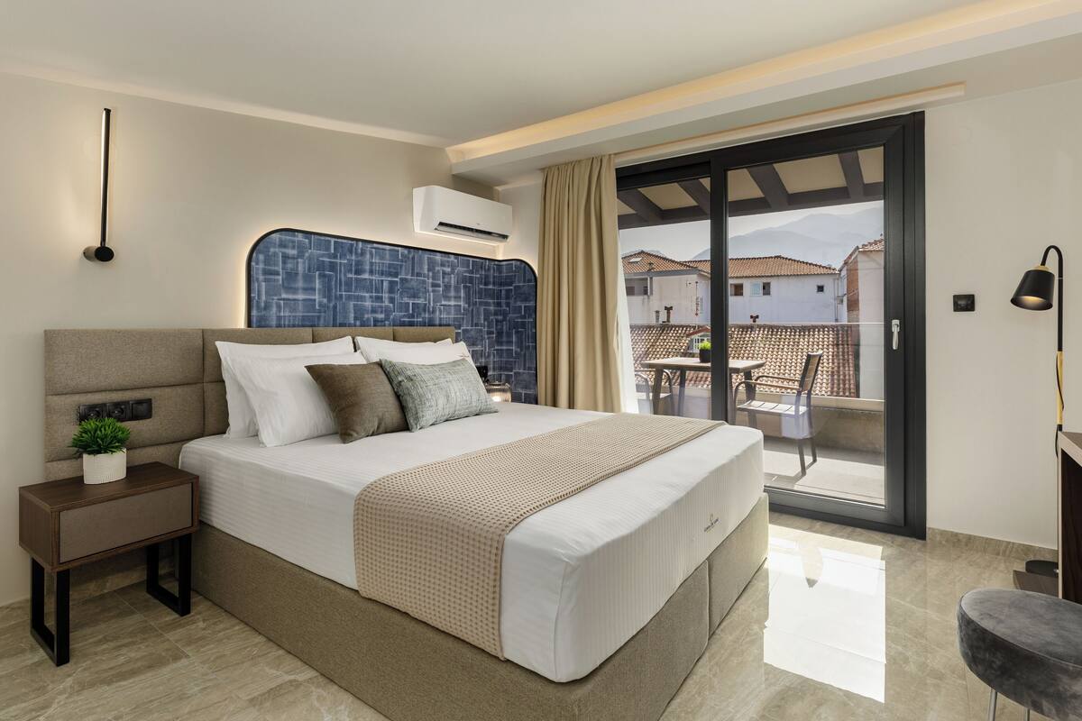A modern bedroom with a large bed, a nightstand with a plant, a blue headboard, and a view of the outside balcony through a glass door.