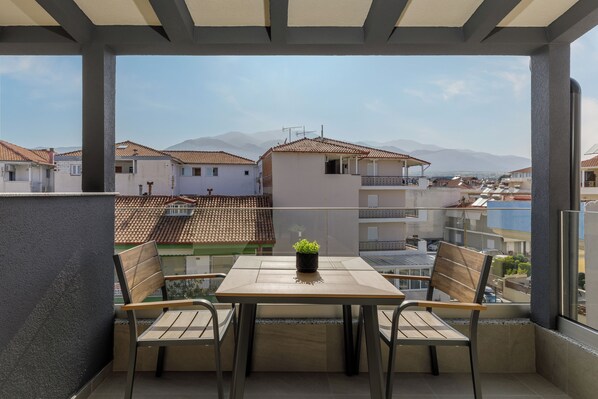 A balcony with a table and chairs, a potted plant, and a view of buildings and mountains. - Costa De Luxe Suites (Dio-Olympos)