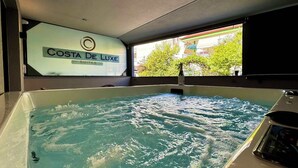 A hot tub with a bottle of champagne, a window with a view of trees and buildings, and a sign reading "Costa De Luxe Suites". - Costa De Luxe Suites (Dio-Olympos)