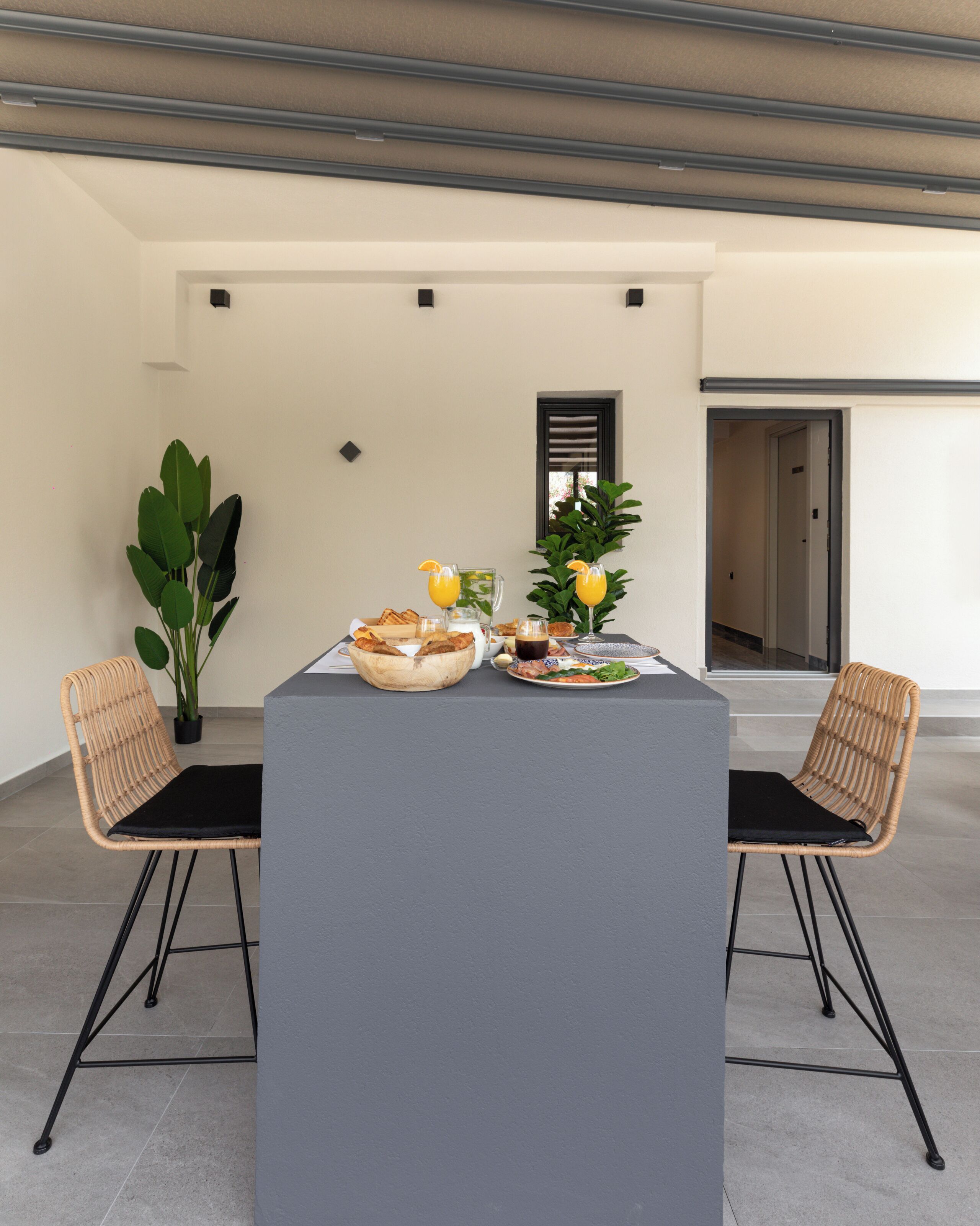 A modern dining area with a central table, two wicker chairs, a potted plant, and a window with blinds.