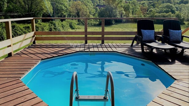 Eine Holzterrasse mit Swimmingpool, zwei Liegestühle mit gemusterten Kissen und Blick auf eine bewaldete Landschaft.