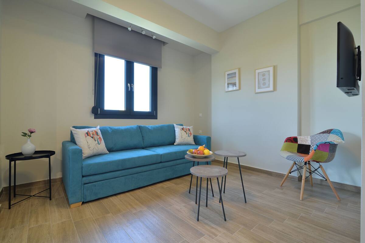 Una sala de estar con un sofá azul, una mesita redonda con fruta, una silla colorida y una ventana con persianas.