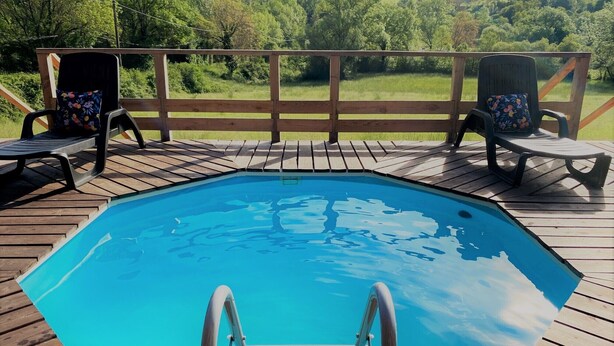 En swimmingpool med træterrasse, to liggestole med blomstrede puder, og et naturskønt udsigt over et grønt landskab.