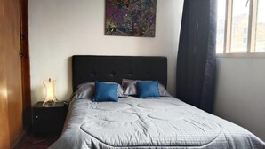 Une chambre à coucher avec un grand lit, une table de chevet avec une lampe, un tableau au mur et une fenêtre avec des rideaux.