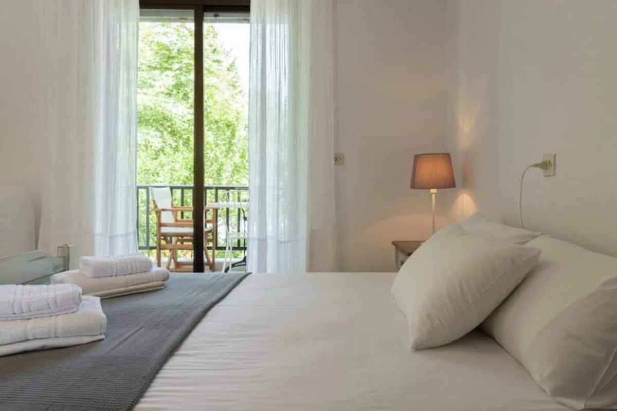 Ein Hotelzimmer mit Bett, Nachttisch, Lampe, einem Fenster mit Vorhängen und Blick ins Freie.