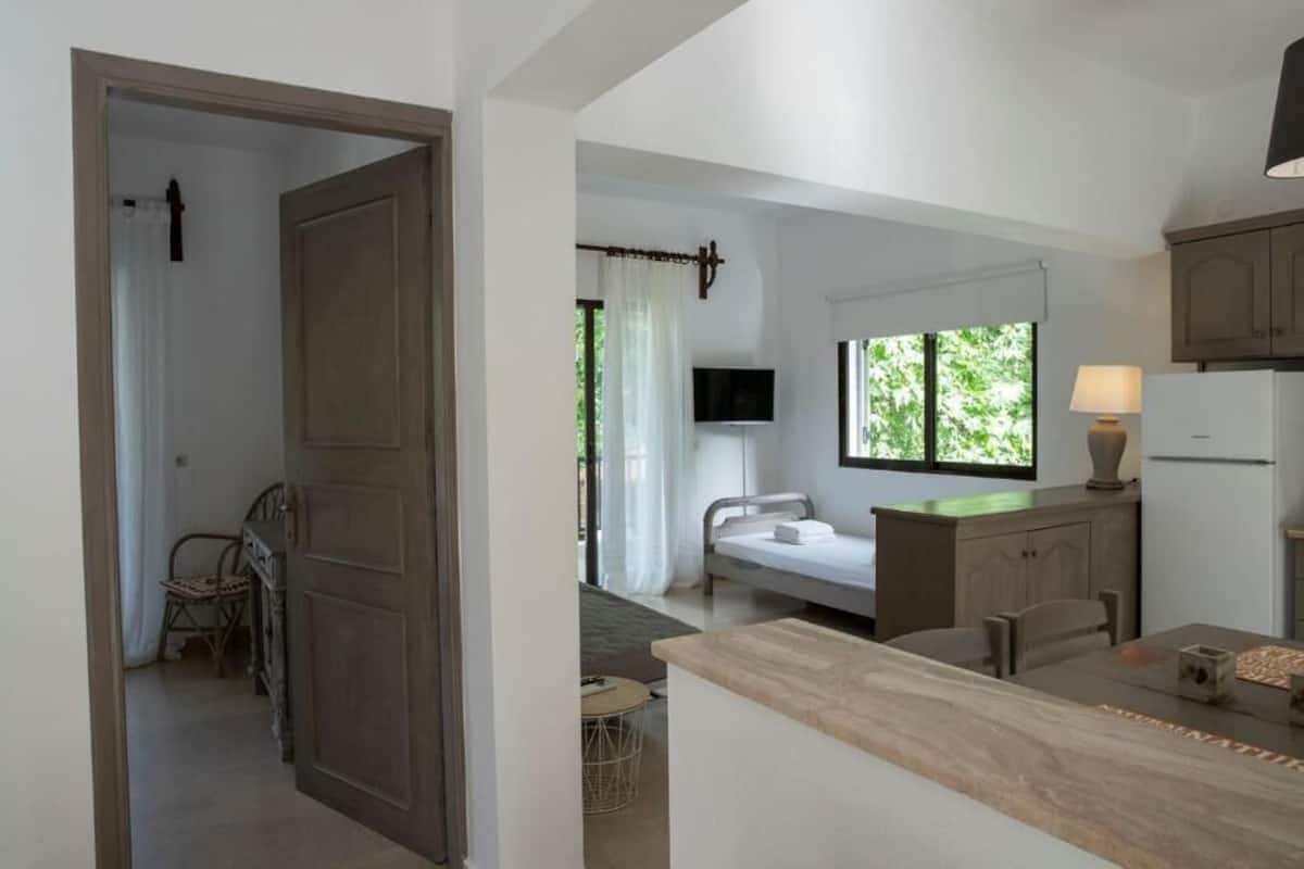Ein modernes Schlafzimmer mit Bett, Nachttisch und Fernseher. Es gibt ein Fenster mit Vorhängen, eine Tür und einen kleinen Tisch mit Stühlen.