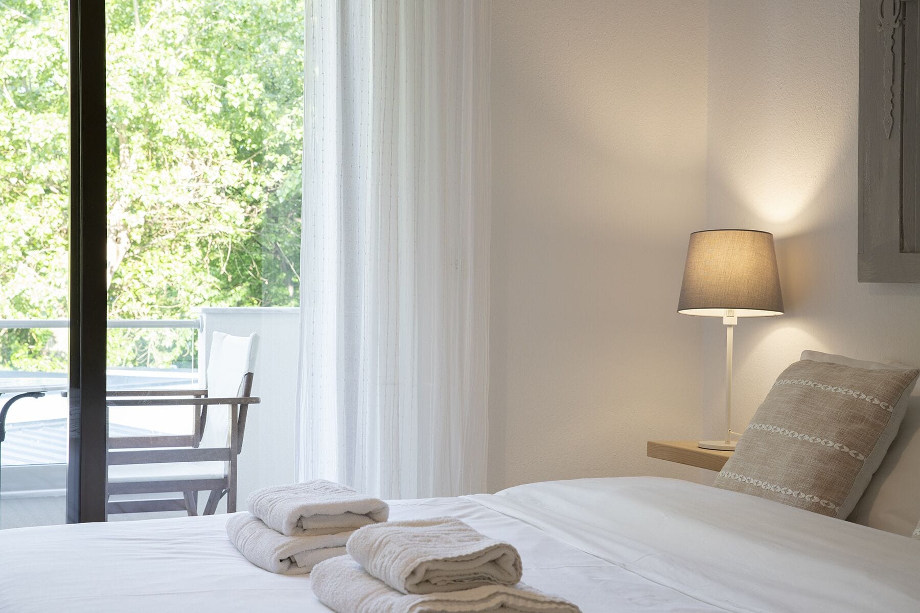 A hotel room with a bed, a lamp, a chair, and a view of greenery outside.
