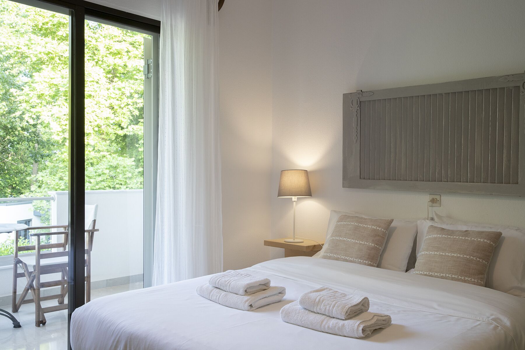 Habitación de hotel con cama, mesita de noche, lámpara y vistas a zonas verdes.