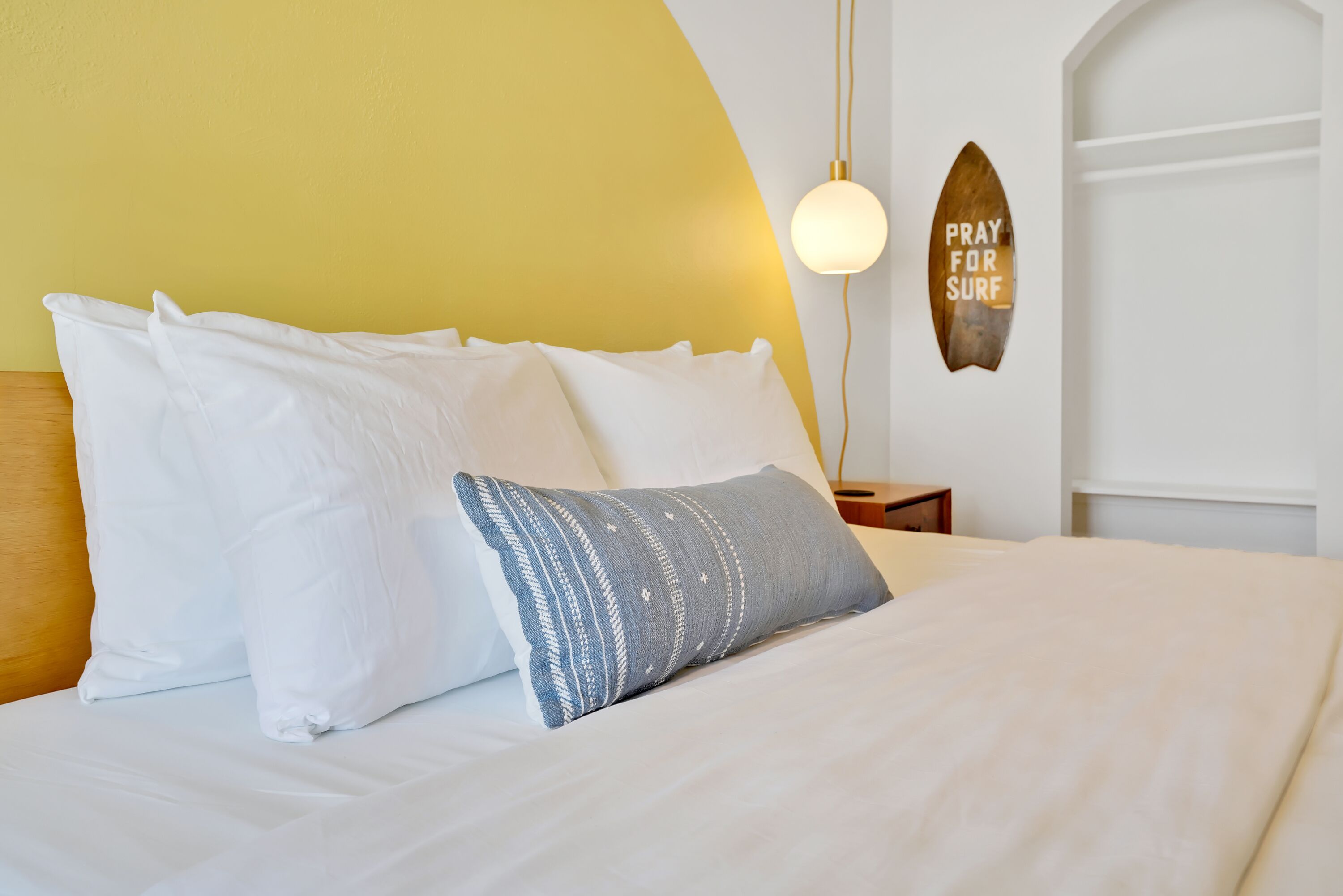 A neatly made bed with white and patterned pillows, a bedside lamp, and a 'PRAY FOR SURF' sign on the wall.