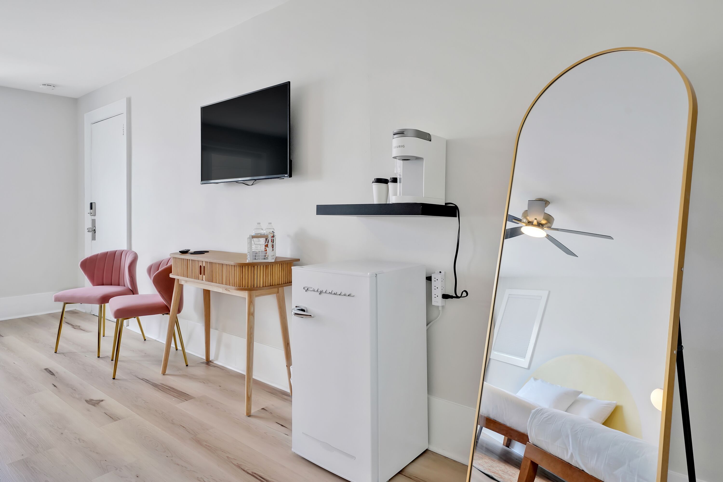 A modern room with a white refrigerator, a wooden table with chairs, a mounted TV, a ceiling fan, and a bed with a white headboard.