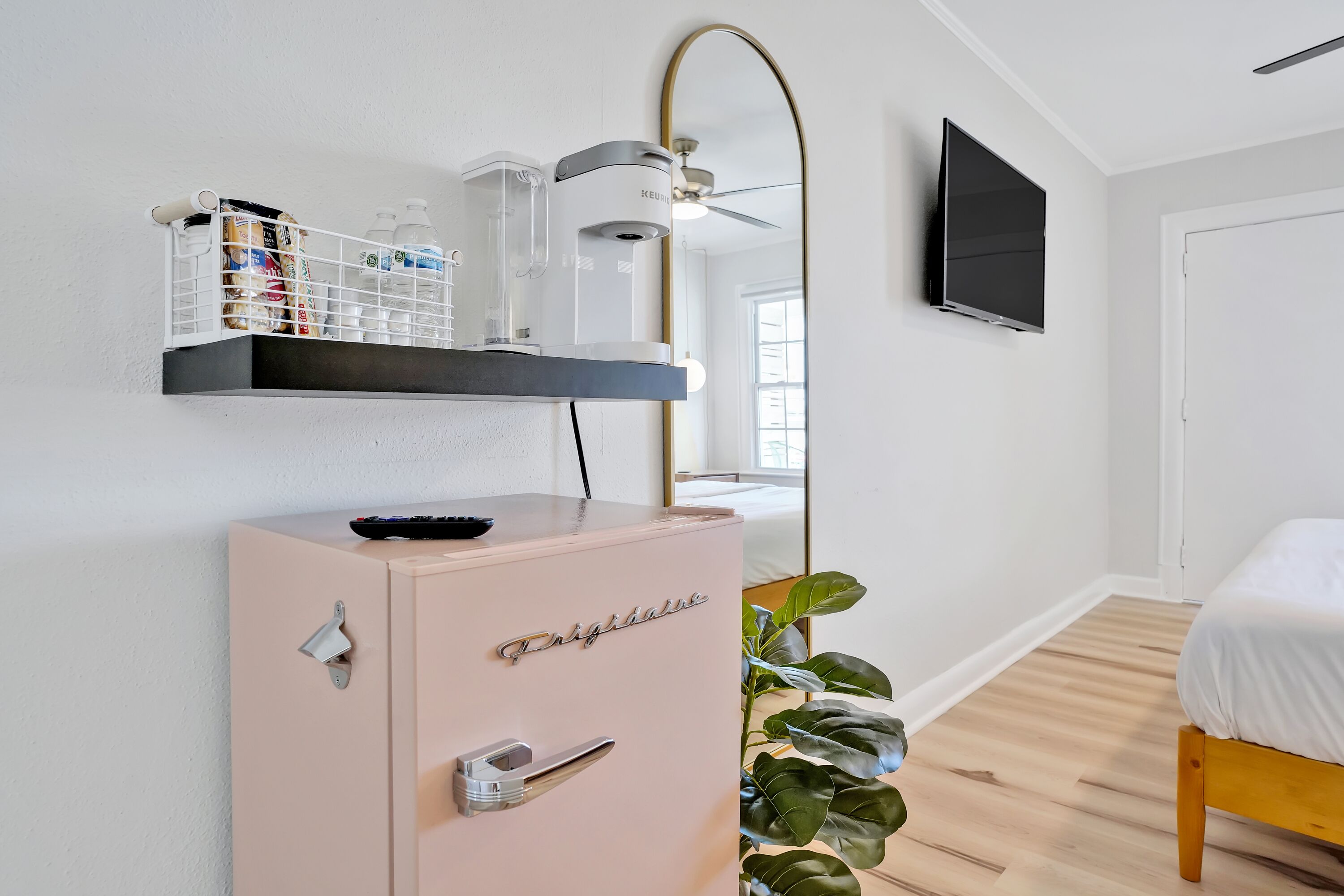 A bedroom with a bed, a TV mounted on the wall, a ceiling fan, a plant, and a minibar.