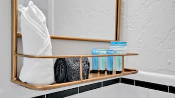 A bathroom with a towel rack holding a white towel and a black towel, a wooden shelf with toiletries, and a mirror.