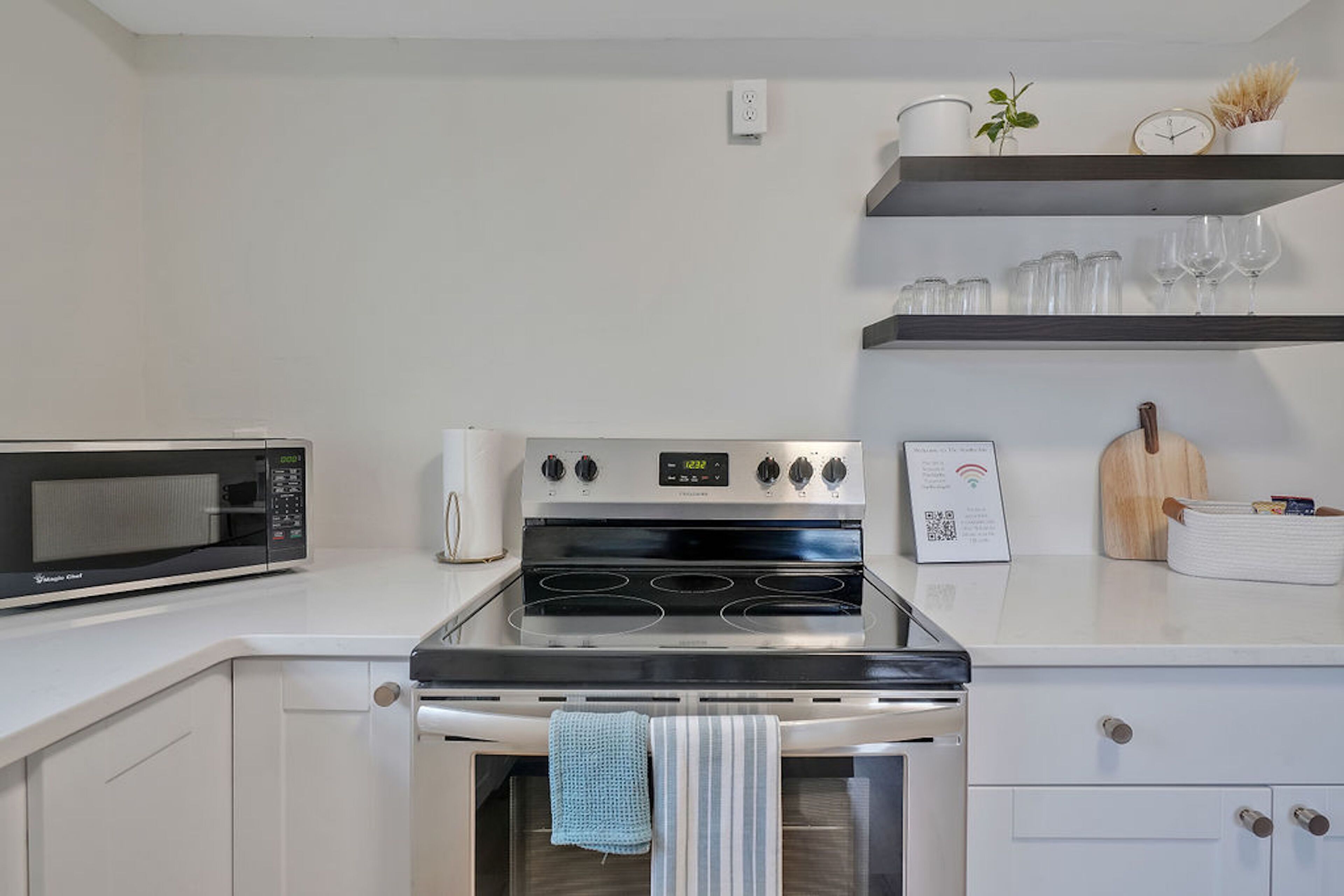 A kitchen with a microwave, stove, oven, and white cabinets. There are shelves above the stove holding glasses and a clock.