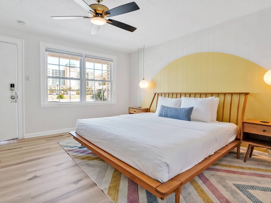 Un dormitorio con cama, cabecera de madera, mesitas de noche, ventilador de techo y ventana con vistas a edificios.