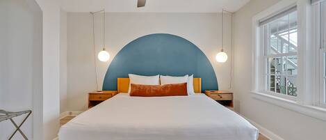A bedroom with a bed, a ceiling fan, two bedside lamps, a window, and a view of a building outside.