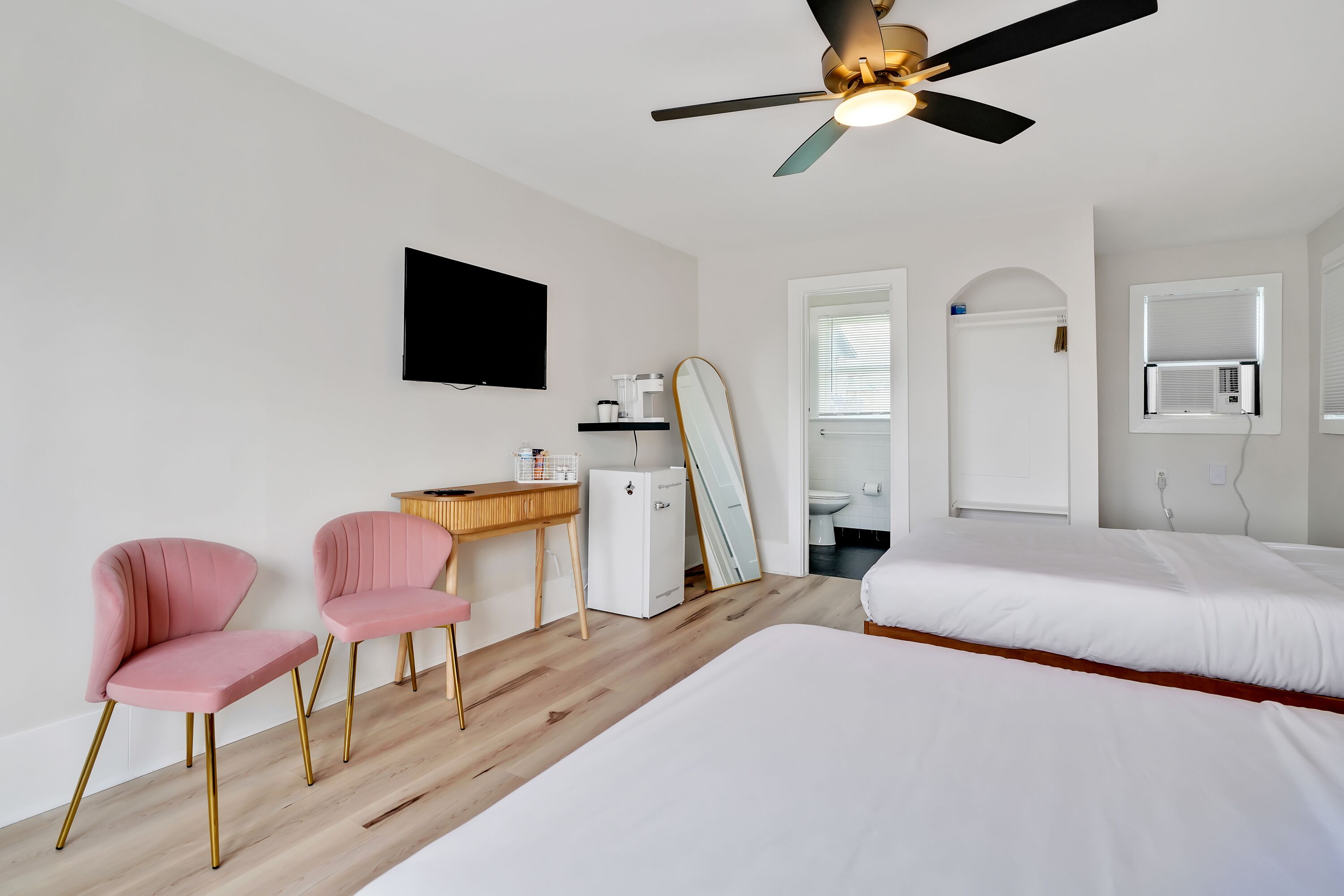 A bedroom with two beds, a desk with a chair, a ceiling fan, and a TV on the wall.