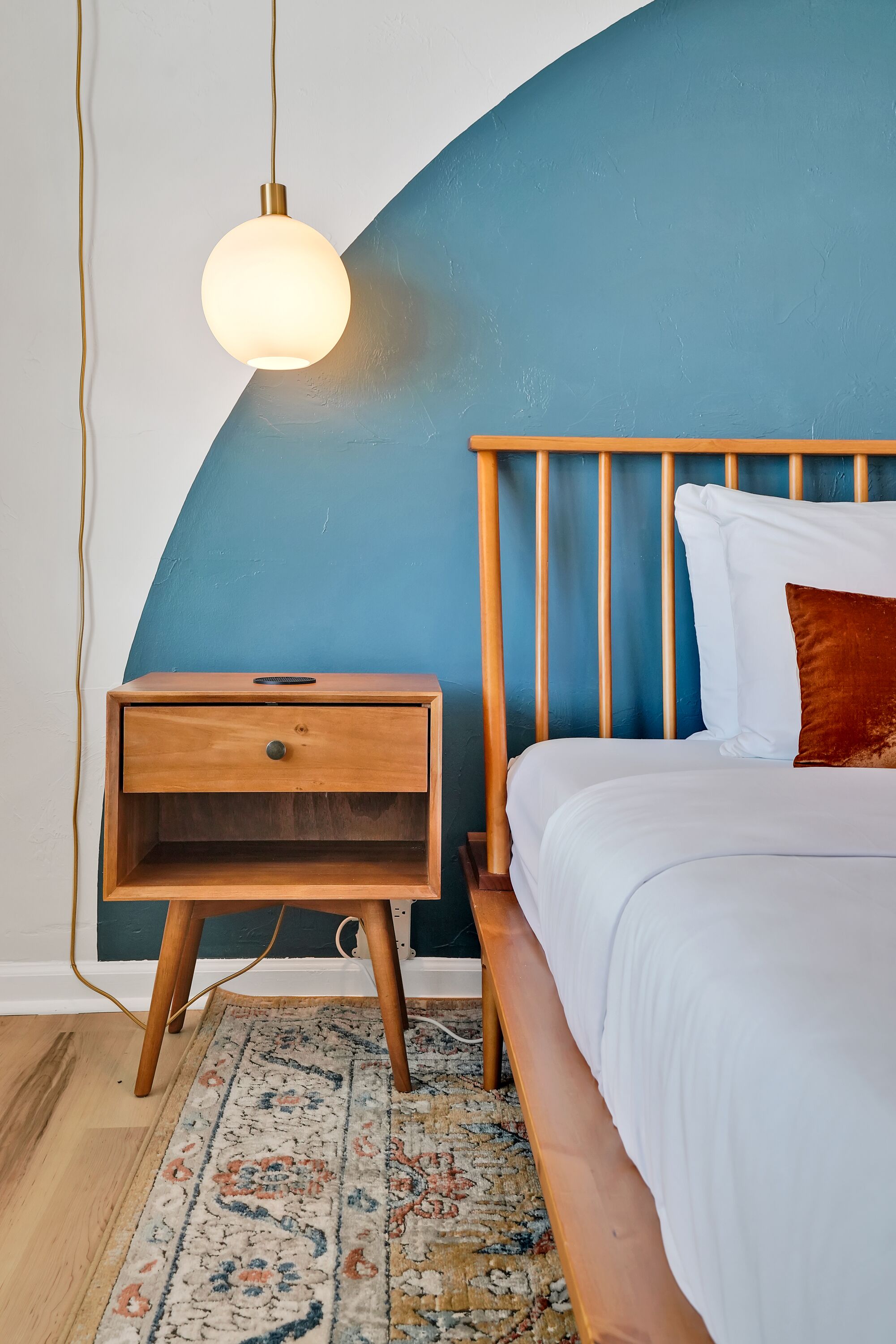 A bedroom with a wooden nightstand, a bed with white bedding, a blue accent wall, and a hanging light fixture.