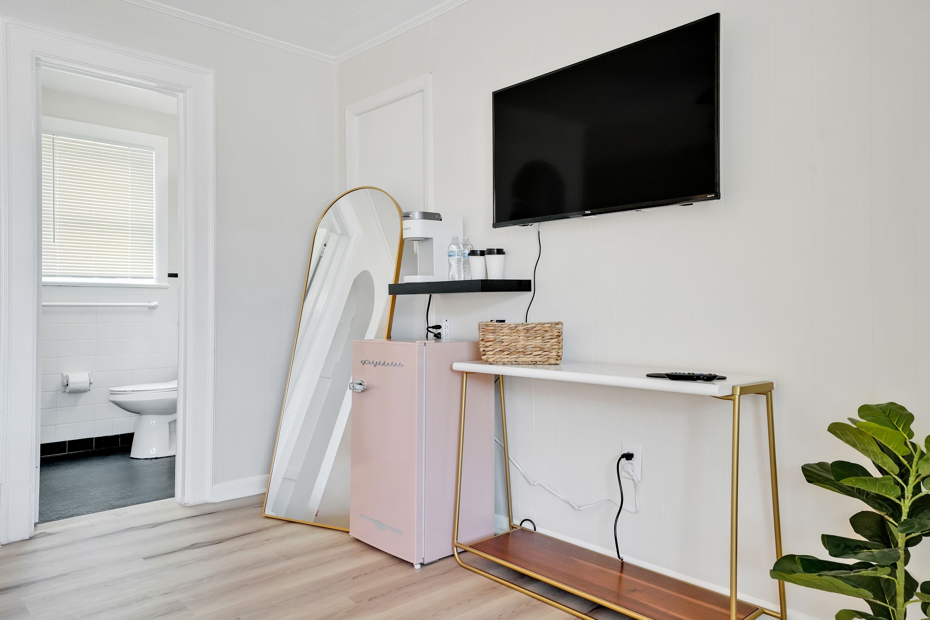 A modern bathroom with a white toilet, a pink refrigerator, a mounted TV, and a wooden floor.