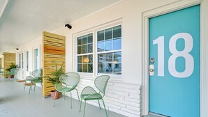 A covered porch with green chairs and potted plants, a teal door with the number 18, and white walls with wooden slats. - The Starlite Inn (Carolina Beach)