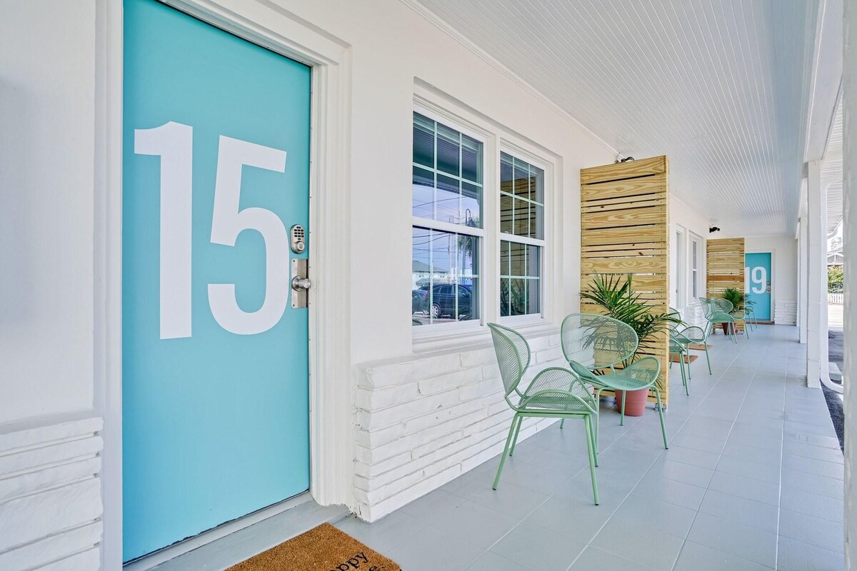 Een gang met een turquoise deur met daarop '15', een witte bakstenen muur, en een rij groene stoelen en tafels.
