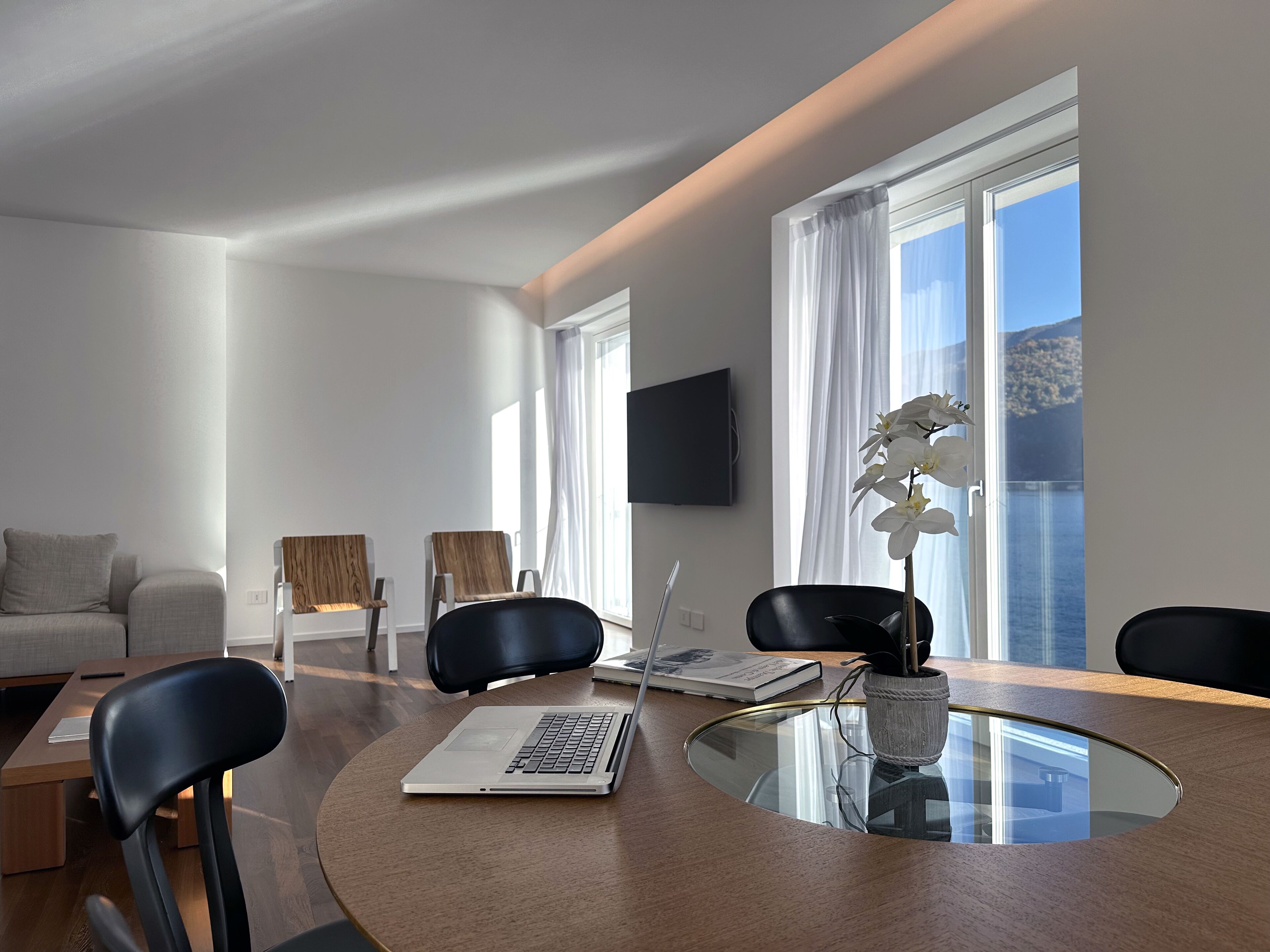 A modern living room with a glass-top dining table, two black chairs, a sofa, and a flat-screen TV mounted on the wall.
