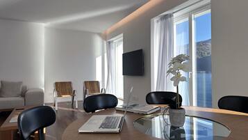 A modern living room with a glass-top dining table, two black chairs, a sofa, and a flat-screen TV mounted on the wall.