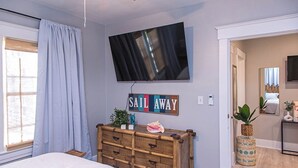 Ett sovrum med en säng, en byrå, en tv som är monterad på väggen och ett fönster med gardiner.