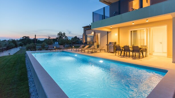 Una piscina moderna al aire libre con sillones de descanso y zona de comedor, rodeada por un césped cuidado y árboles.