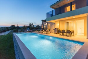 A modern outdoor swimming pool with lounge chairs and a dining area, surrounded by a well-maintained lawn and trees.