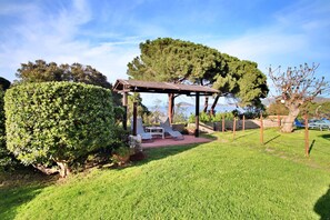 A covered outdoor seating area with lounge chairs, a stone wall, and a view of the sea.